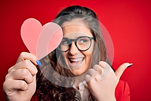 Young beautiful woman with curly hair holding paper heart over isolated red background pointing and showing with thumb up to the