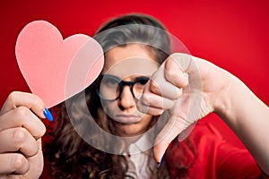 Young beautiful woman with curly hair holding paper heart over  red background with angry face, negative sign showing