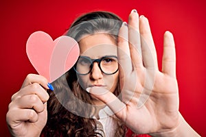 Young beautiful woman with curly hair holding paper heart over  red background with open hand doing stop sign with serious