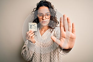 Young beautiful woman with curly hair holding reminder paper with yes word message with open hand doing stop sign with serious and