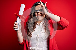 Young beautiful woman with curly hair holding university diploma degree over red background with happy face smiling doing ok sign