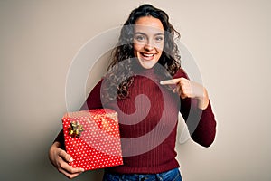 Young beautiful woman with curly hair holding valentine gift over isolated white background with surprise face pointing finger to
