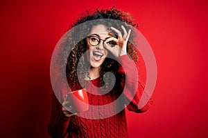 Young beautiful woman with curly hair and piercing drinking red cup of coffee with happy face smiling doing ok sign with hand on