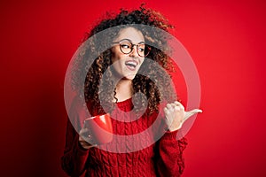 Young beautiful woman with curly hair and piercing drinking red cup of coffee pointing and showing with thumb up to the side with
