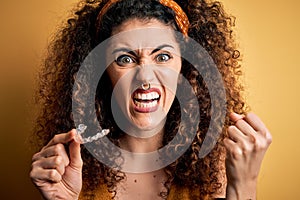 Young beautiful woman with curly hair and piercing holding dental aligner orthodontic annoyed and frustrated shouting with anger,