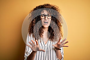 Young beautiful woman with curly hair and piercing wearing striped shirt and glasses afraid and terrified with fear expression