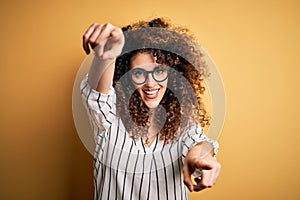 Young beautiful woman with curly hair and piercing wearing striped shirt and glasses pointing to you and the camera with fingers,