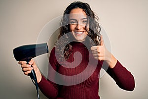 Young beautiful woman with curly hair using hair dryer over isolated white background happy with big smile doing ok sign, thumb up