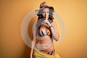 Young beautiful woman with curly hair on vacation wearing bikini and summer hat asking to be quiet with finger on lips