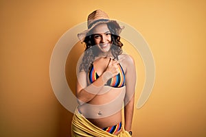 Young beautiful woman with curly hair on vacation wearing bikini and summer hat cheerful with a smile of face pointing with hand