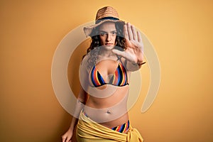 Young beautiful woman with curly hair on vacation wearing bikini and summer hat doing stop sing with palm of the hand