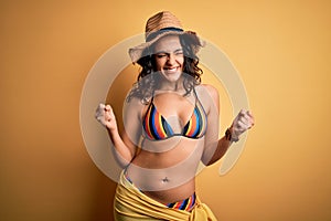 Young beautiful woman with curly hair on vacation wearing bikini and summer hat excited for success with arms raised and eyes