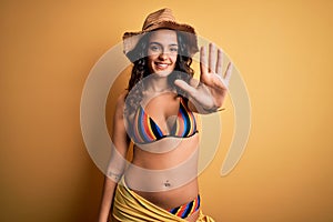 Young beautiful woman with curly hair on vacation wearing bikini and summer hat showing and pointing up with fingers number five
