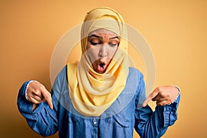Young beautiful woman with curly hair wearing arab traditional hijab over yellow background Pointing down with fingers showing