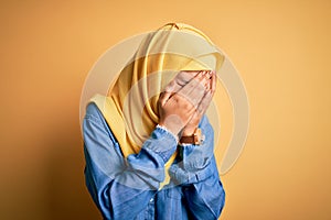 Young beautiful woman with curly hair wearing arab traditional hijab over yellow background with sad expression covering face with