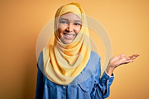 Young beautiful woman with curly hair wearing arab traditional hijab over yellow background smiling cheerful presenting and