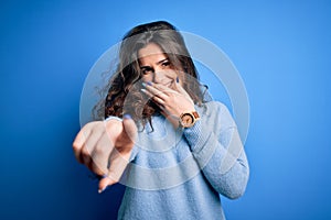 Young beautiful woman with curly hair wearing blue casual sweater over  background laughing at you, pointing finger to the