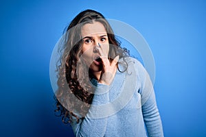 Young beautiful woman with curly hair wearing blue casual sweater over isolated background hand on mouth telling secret rumor,