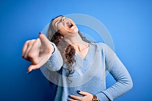 Young beautiful woman with curly hair wearing blue casual sweater over isolated background laughing at you, pointing finger to the
