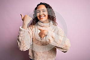 Young beautiful woman with curly hair wearing casual sweater standing over pink background Pointing to the back behind with hand