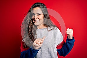 Young beautiful woman with curly hair wearing casual sweatshirt over  red background Pointing to the back behind with hand