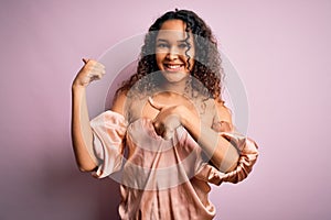 Young beautiful woman with curly hair wearing casual t-shirt standing over pink background Pointing to the back behind with hand