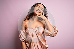 Young beautiful woman with curly hair wearing casual t-shirt standing over pink background smiling doing phone gesture with hand