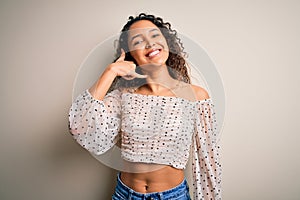 Young beautiful woman with curly hair wearing casual t-shirt standing over white background smiling doing phone gesture with hand