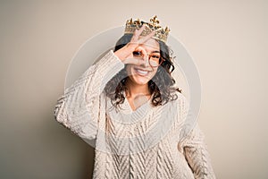 Young beautiful woman with curly hair wearing golden queen crown over white background doing ok gesture with hand smiling, eye