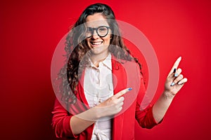 Young beautiful woman with curly hair wearing jacket and glasses over red background smiling and looking at the camera pointing