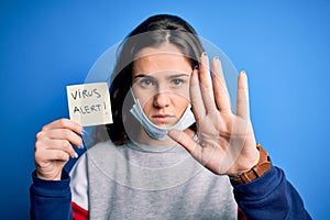 Young beautiful woman with curly hair wearing mask holding reminder with virus alert message with open hand doing stop sign with
