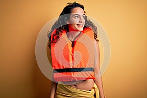 Young beautiful woman with curly hair wearing orange lifejacket over yellow background looking away to side with smile on face,