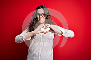 Young beautiful woman with curly hair wearing shirt and glasses over red background smiling in love showing heart symbol and shape