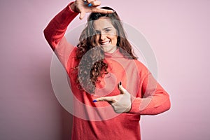 Young beautiful woman with curly hair wearing turtleneck sweater over pink background smiling making frame with hands and fingers