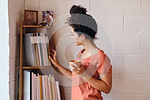 Young beautiful woman with dark curly hair in T-shirt holding cup of coffee in hand dreamily choosing book from