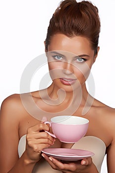 Young beautiful woman with dark hair picked up holding a ceramic cup and saucer pale pink drink tea or coffee on a white back