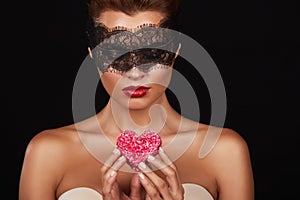 Young beautiful woman with dark lace on eyes bare shoulders and neck, holding cake shape of heart to enjoy the taste and are