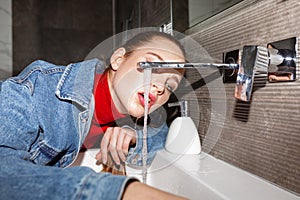 Young beautiful woman in denim jacket drinking tap water from sink