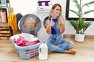 Young beautiful woman doing laundry sitting by wicker basket pointing fingers to camera with happy and funny face