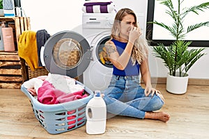 Young beautiful woman doing laundry sitting by wicker basket smelling something stinky and disgusting, intolerable smell, holding