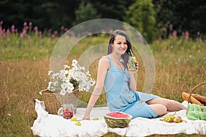 A young beautiful woman in dress is drinking lemonade from glass jar, sitting on plaid on green grass. Picnic basket