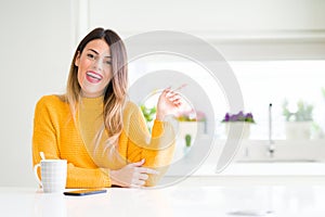 Young beautiful woman drinking a cup of coffee at home with a big smile on face, pointing with hand and finger to the side looking