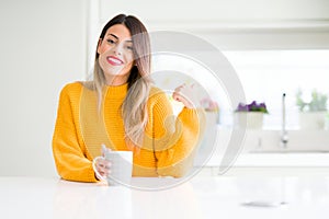 Young beautiful woman drinking a cup of coffee at home pointing and showing with thumb up to the side with happy face smiling