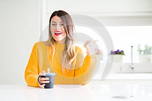 Young beautiful woman drinking a cup of coffee at home pointing and showing with thumb up to the side with happy face smiling