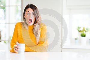 Young beautiful woman drinking a cup of coffee at home scared in shock with a surprise face, afraid and excited with fear