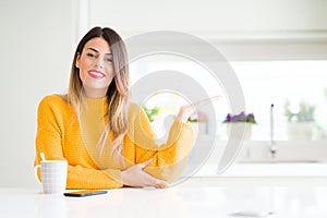 Young beautiful woman drinking a cup of coffee at home smiling cheerful presenting and pointing with palm of hand looking at the