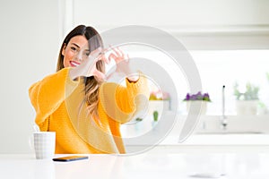 Young beautiful woman drinking a cup of coffee at home smiling in love showing heart symbol and shape with hands
