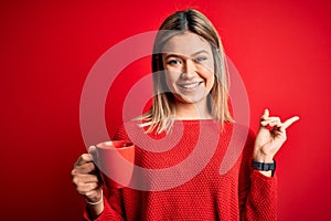 Young beautiful woman drinking cup of coffee standing over isolated red background very happy pointing with hand and finger to the