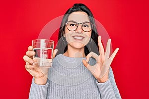 Young beautiful woman drinking a glass of fresh water over isolated red background doing ok sign with fingers, excellent symbol