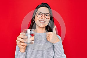 Young beautiful woman drinking a glass of fresh water over isolated red background happy with big smile doing ok sign, thumb up